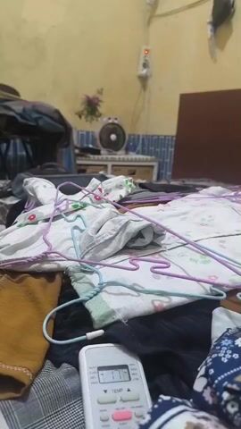 The video shows a messy bedroom, possibly a dorm room, with clothes strewn across the bed and floor. There are several hangers on the bed, some holding clothes. A remote control for an air conditioner is visible in the foreground, showing a temperature of 17 degrees Celsius. In the background, there is a fan, a dresser, and a wardrobe. The walls are a pale yellow color. There are no people visible in the video. The lighting is dim, suggesting it may be nighttime or the room is not well-lit. The overall impression is one of disarray and clutter.