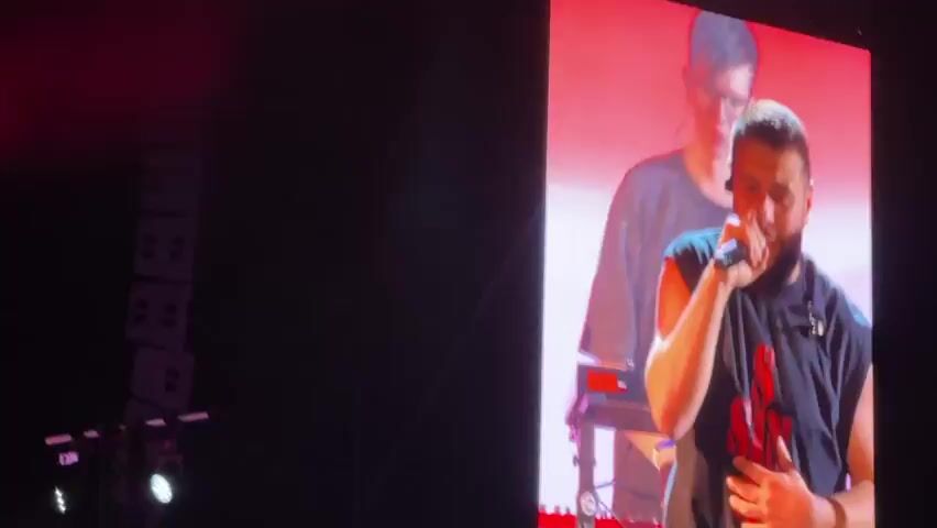 The video shows a live musical performance on a stage with a large pink screen in the background displaying a circular design. On the right side of the stage, a male singer in a sleeveless blue shirt and white pants is holding a microphone to his mouth with his right hand and gesturing with his left hand. Behind him, another person is partially visible. To the left of the singer, there are other band members. One person in a black t-shirt is playing a guitar. Another person in a gray t-shirt is operating a keyboard. Further to the left, another person in a black t-shirt is standing, possibly playing an instrument or singing. The stage is lit with red and purple lights, and there is some stage smoke. On the far left of the stage, there are stacks of speakers and lighting equipment. The audience is not clearly visible, but the perspective suggests the viewer is among them.