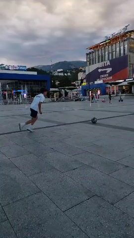 The video shows a young person in a public square playing with a soccer ball. The person is wearing a white t-shirt and blue shorts. They are kicking the ball and running after it. The square is paved with large square tiles and there are buildings and trees in the background. The sky is cloudy and it appears to be dusk. There are other people in the background, but they are not interacting with the person playing soccer.