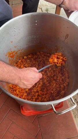 A man's hand reaches into a large pot of pasta with tomato sauce. The pot is made of shiny metal with two handles and is sitting on a red plastic tray on a brick surface. The pasta appears to be ditalini. The man scoops pasta with a spoon in his right hand, then uses a fork in his left hand to stir the pasta. A second person, partially visible, is standing nearby.