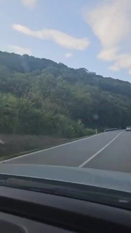 A first-person perspective video shows a car driving on a paved road. The road is curving to the right. The right side of the road is bordered by a steep, tree-covered hill. The left side of the road has a metal guardrail and more trees and vegetation. The sky above is blue with scattered white clouds. The sun is shining, and there are lens flares visible on the camera. The interior of the car is visible in the foreground, including the dashboard and windshield. No other people or vehicles are clearly visible.