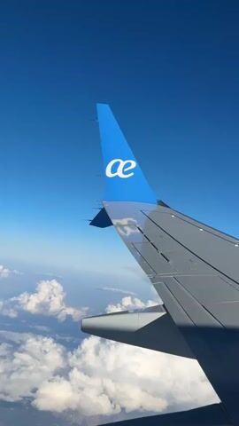 The video shows a view from an airplane window. The plane's wing is visible in the foreground with a blue tail fin displaying a white logo that resembles the lowercase letters "ae" in a stylized font. Below the wing are fluffy white clouds. Above, there is a clear, bright blue sky. As the video progresses, the moon comes into view in the upper center of the frame.