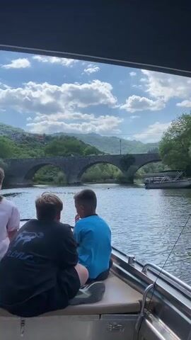 Three individuals, two boys and a girl, are seated on a boat, facing away from the camera. They are looking towards a picturesque landscape featuring a river and a stone bridge with multiple arches. The bridge spans across the river, and trees line the banks. In the background, lush green hills rise under a partly cloudy blue sky. The sun appears to be shining, suggesting it's daytime, possibly late morning or afternoon. The overall mood seems peaceful and scenic, indicative of a boat trip on a pleasant day. The scene is identified as Rijeka Crnojevića, Montenegro. On the back of the black t-shirt worn by the boy in the middle, there is visible text, though it is partially obscured and difficult to read clearly, but it appears to say "BRONCO".