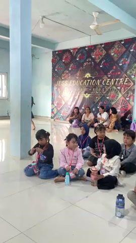 This video shows a group of young children sitting on the floor in a room. A banner in the background reads "JESS EDUCATION CENTRE". The children are facing a group of adults who are also sitting. Some of the children are holding water bottles. The room appears to be a classroom or community center.