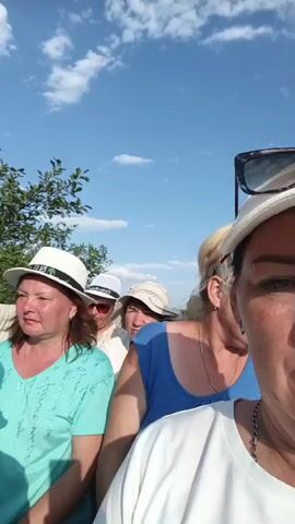 The video shows a group of people, mostly women, wearing hats and looking at the camera. The person filming is speaking and addressing the group, asking them to be careful. The environment appears to be outdoors with trees and a blue sky with clouds in the background. The people are close together, suggesting they are on a vehicle or in a crowded space. The overall mood seems lighthearted and possibly humorous, as one woman is laughing.