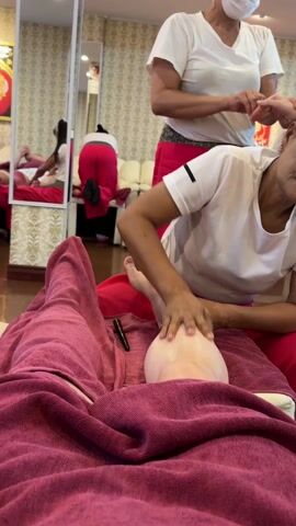 A person is receiving a leg massage from a masseuse in a spa. The person receiving the massage is lying on a massage table covered with a burgundy-colored towel, with their lower leg exposed. The masseuse, wearing a white t-shirt and red pants, is applying oil and massaging the person's calf and foot. Another masseuse, also in a white t-shirt and red pants, is visible in the background, standing and attending to another customer whose back is visible in the mirror. The spa has tiled floors and decorated wallpaper. Other massage tables and a white sofa are also visible in the background, and a black pen lies on the towel next to the person's leg. The lighting is bright and even.