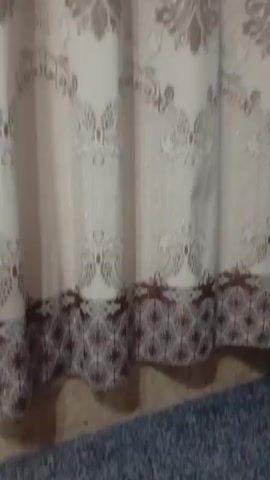 A white floor fan is shown next to a curtain. The fan has a digital display on its base that shows numbers. The curtain has a patterned design. The lighting is dim.
