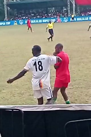 The video depicts a soccer game on a grassy field, seen from behind a barrier, likely a metal railing. A player wearing a white jersey with the number 13 on the back walks along the sideline, accompanied by a player in a red uniform. A referee in a yellow shirt is visible on the field further away. The background shows spectators in the stands, and signage related to the "CLEB MALALAN SUPER CUP." An individual in a striped shirt is positioned near the barrier, possibly part of the event staff. The overall scene captures a moment during the soccer game, possibly a player substitution or a break in the action.