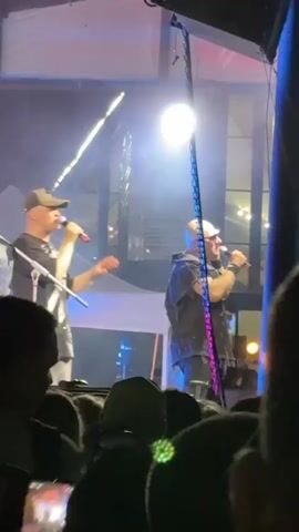 A video captures a live music performance on an outdoor stage at night. Two men are on stage singing into microphones. The man on the left wears a cap and a black and white checkered shirt, holding a microphone and gesturing with his other hand. The man on the right wears a dark shirt and also holds a microphone, facing the audience. A large bright light is suspended above the stage, and stage lighting casts blue and white beams. Behind the stage, a large sign with the text "90-00" in colorful letters is visible. The stage is partially covered by a tent-like structure. In the foreground and background, a crowd of people watches the performance. Many audience members are holding up their phones to record or take pictures. The environment appears to be a festival or concert venue, with trees visible in the background and a tower-like structure with lights in the distance. The overall atmosphere is energetic and celebratory.