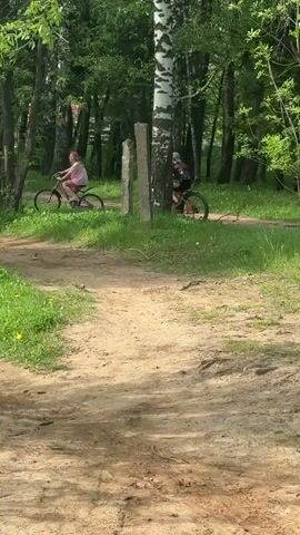 The video depicts three individuals riding bicycles on a dirt path through a wooded area. The path is flanked by grass and scattered dandelions. Trees, including birch trees, line the sides of the path. The camera remains stationary, focusing on the cyclists as they move away from the viewer. The sky is not visible, and the lighting suggests it is daytime.