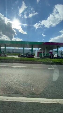 A gas station with multiple cars lined up at pumps is visible under a bright, sunny sky with fluffy clouds. The perspective is from inside a vehicle, looking out the side window. The road in the foreground is asphalt with white lines. The gas station canopy is white and pink, and a tall sign with green and white branding stands to the right. Several cars are parked and queued at the pumps. The environment appears to be a rural or suburban area with grassy fields and some distant buildings. The camera is moving, suggesting the video was recorded from a moving vehicle.