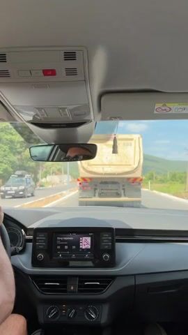 The video shows the interior of a car from the perspective of the driver or front passenger. In the foreground, the dashboard of the car is visible, including the steering wheel on the left, the central infotainment screen displaying a music player interface, and climate control buttons. The rear-view mirror is also visible, reflecting the driver's head and arm. Outside the car, a yellow dump truck is driving on a road ahead. The road appears to be under construction, with barriers and unfinished surfaces visible. Trees and hills form the background under a clear sky. The overall scene suggests a moment of travel on a sunny day.