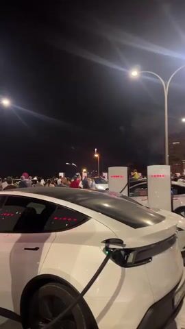 The video captures a nighttime scene with several white Tesla vehicles parked and charging at charging stations. A crowd of people is gathered in the background, some appearing to be looking at fireworks in the sky. The atmosphere is lively, with artificial lighting from streetlights and the charging stations illuminating the area. The focus is on the electric vehicles and the charging infrastructure, with the crowd and fireworks serving as a backdrop to the scene.