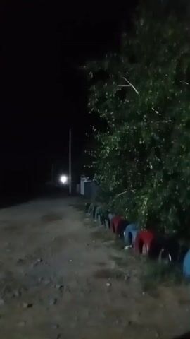 The video shows a dimly lit outdoor scene at night. A dirt path runs through the frame, bordered on the right by a row of colorful tires, red and blue, possibly used as a barrier or decoration. Behind the tires, there is dense foliage from a large tree. A utility pole is visible further down the path, with a faint light emanating from its base. To the left of the path, the scene is mostly dark, with some indistinct structures. The ground appears to be rough and uneven. The camera is moving, suggesting it is handheld or mounted on a moving vehicle. The overall atmosphere is dark and quiet.