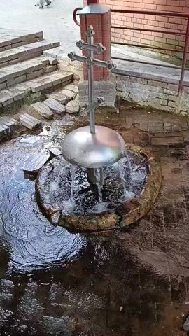 A video shows a spring with water flowing out from under a large silver dome. A tall metal cross stands in the center of the spring. The spring is surrounded by a circular stone border. In the background, there are stone steps leading up to a brick wall. A person is partially visible walking up the steps. The ground around the spring is wet and appears to be tiled.