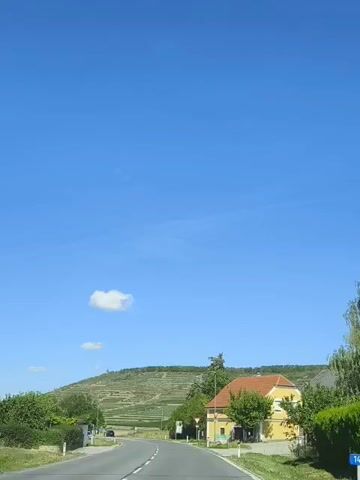 A rural road curves to the left, leading past a yellow building with an orange roof. To the left of the road, there's a small white building and several parked cars. On the right side of the road, a yield sign is visible. In the background, a green hill slopes upwards, covered in terraced fields. The sky is a clear, bright blue with a single small cloud.