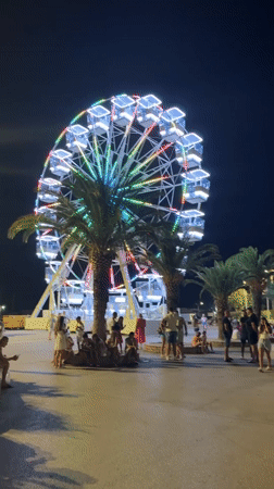 Bar's waterfront night fair: Ferris wheel, market stalls, and street sculptures.
