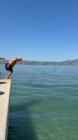 A man in dark shorts jumps off a concrete dock into clear green water under a bright blue sky. Mountains are visible in the background. He enters the water with a splash and then submerges, creating a circular ripple on the surface. He then surfaces and swims, visible as a head and shoulders in the water, with more ripples spreading around him.