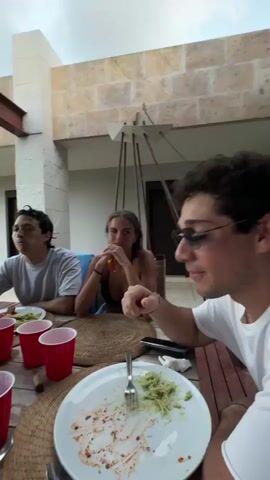 The camera pans around a partially eaten meal at a wooden table, where a group of young adults are dining on an outdoor patio. Several red plastic cups are scattered around the table, along with plates of food. A man in a black t-shirt and baseball cap stands behind the table, seemingly interacting with the group. The patio is part of a large house or resort, with a thatched roof structure visible in the background. The sky is overcast.