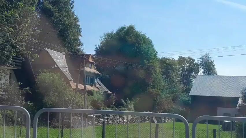 The video shows a view from a vehicle looking out at a rural landscape on a sunny day. In the foreground, there are two metal railings. Beyond the railings is a grassy area and a stone wall. To the left, there is a house with solar panels on its roof, partially obscured by trees. To the right, there is a dark-colored building with a white roof. Lush green trees and foliage are visible throughout the scene, and the sky is a clear blue. There are no people or animals visible in the frame, and no actions or interactions are occurring. The overall impression is one of a quiet, suburban or rural setting.