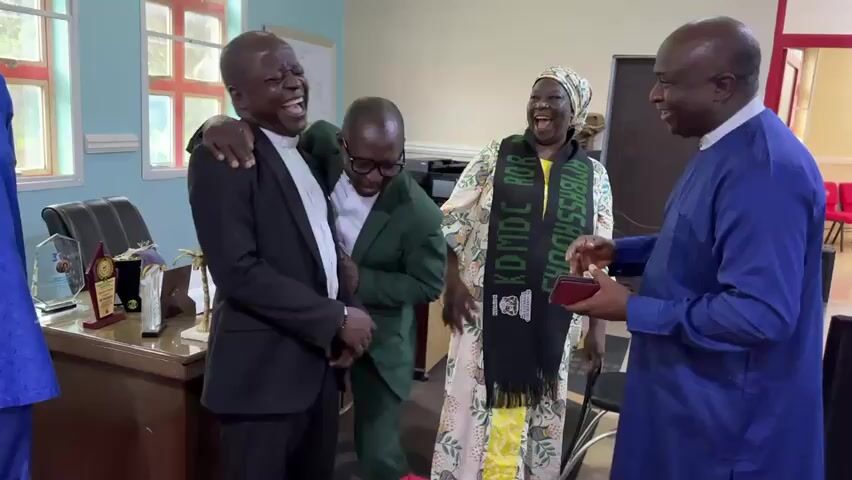 A group of five men and one woman are gathered in an office. The woman is wearing a headscarf and a patterned dress with a black scarf draped around her neck that says "KDMC". One man in a black suit is laughing and has his arm around another man in a green suit, who is also laughing and wearing glasses. A man in a blue shirt is holding a phone and appears to be speaking to the group. Another man in a blue shirt and pants is on the far left, with his hand raised. A man in a light brown outfit is standing to the left of the man in the blue shirt. Behind the laughing men is a desk with trophies on it and a printer. The room has light blue walls and windows with red frames.