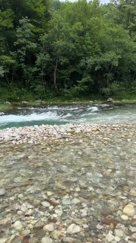The video captures a scenic riverside location, likely in Džajići, Bosnia and Herzegovina, with a focus on outdoor recreational activities. The scene opens with a view of a clear, shallow river flowing over a bed of smooth stones, with lush green trees lining the opposite bank and a gentle slope of the terrain behind.

The central figures are a young woman and a child, both equipped for water activities. The woman, wearing a helmet and a life vest, is seen taking a selfie in the first part of the video, with a blue van and a yellow inflatable raft visible in the background. The setting appears to be a riverbank, strewn with pebbles, with a building nestled on a hillside behind her. Later, the child, also in a life vest and a pink skirt, is filmed in a blurry shot, standing by the river and smiling. The overall impression is one of a pleasant, possibly adventurous, day spent by the water, with a sense of fun and excitement. The weather appears overcast, indicated by the bright, diffused light. The presence of the raft suggests an activity like rafting or kayaking is planned or has just concluded. The location cues, such as the style of the building and the natural landscape, are consistent with the described location.