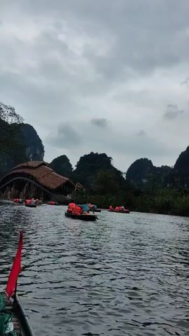 The video shows a body of water with boats navigating towards a structure with a thatched roof. The structure appears to be built over the water, potentially a bridge or a building connected by a bridge. People in orange life vests are visible in the boats. The sky is overcast and the surrounding landscape includes lush greenery and mountains in the background. The camera appears to be positioned on a boat, giving a perspective of moving on the water towards the thatched-roof structure. A red flag is visible at the front of the boat on which the video is being recorded.