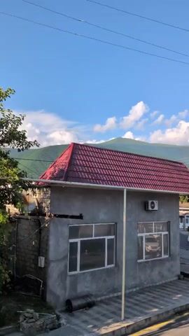 This media file captures a daytime scene in a rural or semi-rural setting, likely in Şəki, Azerbaijan, given the location context. The primary focus is a small, grey concrete building with a prominent red tiled roof. The building appears to be a commercial or utilitarian structure, with large, white-framed windows. One window on the right side of the building shows the interior with a parked vehicle, possibly a car or van, visible. An air conditioning unit is mounted on the exterior wall above this window.

To the left of the building, there is a section of brick wall, and beyond that, lush green foliage from a tree. The ground in front of the building is paved with interlocking stones and a yellow line demarcates the edge of a road or pathway. A white pipe runs vertically along the front of the building, and a black rolled-up material, possibly tarpaulin or roofing felt, lies on the ground near the left window.

In the background, rolling green hills and mountains dominate the landscape, suggesting a location with natural beauty. The sky is a vibrant blue with scattered white clouds, indicating fair weather and likely midday or early afternoon. There is no visible text within the media file itself. The overall impression is one of a simple, functional building situated within a scenic, natural environment under a pleasant sky.