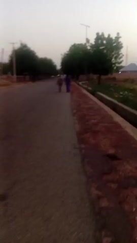 A somewhat blurry, low-light video shows a paved road with a raised concrete walkway on the right. A few trees are visible, along with some buildings and a tall pole with a light fixture. Two figures, one in a purple garment and another in a red garment, are walking away from the camera on the road. The sky is a pale, hazy grey, suggesting it is either early morning or late evening. There is some evidence of vegetation on the right side of the road. The video appears to be shot from a moving perspective, possibly from a vehicle or by someone walking.