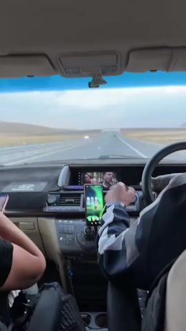 The video shows the interior of a car from the perspective of a passenger. The car is in motion, driving on a highway. A driver, wearing a blue and white jacket, is visible on the right side, holding the steering wheel. A second person, whose arm is visible on the left, is also in the car. A smartphone is mounted on the dashboard, displaying a green app icon. The car's infotainment screen is on, showing two people. The dashboard is dark brown, and the car's interior appears to be a neutral color. The road ahead is straight, with a clear sky and barren landscape on either side.