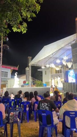 The video shows a fashion show at night in an outdoor venue. A woman in a long, light-colored, tiered gown is walking on a stage. A crowd of people is sitting in blue plastic chairs facing the stage. The stage has a white tent-like covering with chandeliers hanging beneath it. There are speakers stacked on the side of the stage, and a backdrop with a logo that says "Samui Group." Behind the stage, there are buildings, including a traditional Thai building with a red roof. The sky is dark, and there are trees visible on the left side of the frame. There are spotlights on the stage illuminating the model.