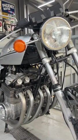 The video shows a close-up panning shot of a vintage silver Benelli 750 Sei motorcycle. The camera focuses on the engine, the silver fuel tank with the Benelli logo, the black textured seat, and the rear suspension with red springs. The motorcycle is stationary, likely in a garage or showroom, with posters and shelves visible in the background. The lighting is bright, highlighting the motorcycle's chrome and silver components.