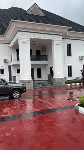 A grey SUV is parked on a wet, red-paved surface in front of a large white building with a black roof. The building has large white columns supporting an upper-level balcony. Two individuals, dressed in dark clothing, are exiting the SUV. One individual appears to be handing a rifle to another individual. Another person with a camera is visible to the right of the SUV, possibly filming the event. The sky is overcast.