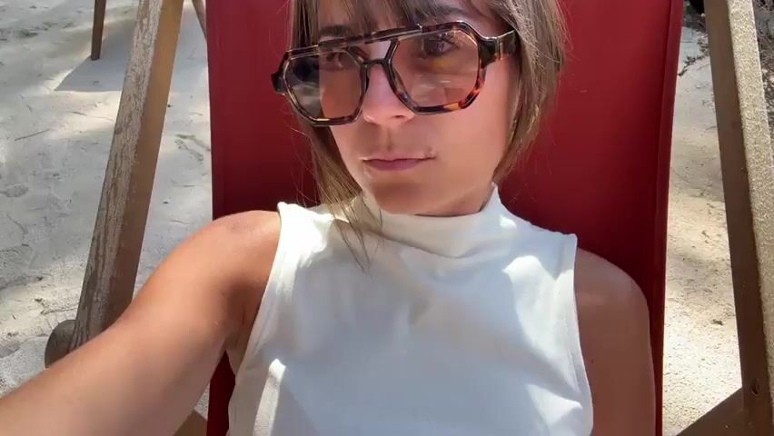 A woman wearing sunglasses and a white sleeveless top is sitting in a red and wood deck chair. She also has on striped pants. The chair is placed outdoors on a concrete surface that appears to be in the shade.  A small portion of foliage is visible in the background. The camera's vantage point is slightly above and to the side of the woman, capturing a view from her chest down to her lap. Background music plays.