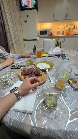 The video shows a kitchen and dining area. A table covered with a grey and white patterned tablecloth is set for a meal with plates of food, glasses, and drinks. There are cigarettes and a lighter on the table.  A person's hand with a silver watch is visible, holding a cigarette. In the background, a television is showing a news program. To the side of the table, there's a JBL party speaker with green lights on.  A jacket is draped over one of the chairs. The kitchen area in the background has white cabinets and appliances. There's an oven, microwave, and a refrigerator.  The overall lighting is somewhat dim.