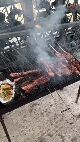 The video shows a close-up of a barbecue grill with various meats cooking over charcoal. Ribs are arranged on the left side of the grill, some of them already showing a grilled texture and smoke rising. To the right of the ribs, more ribs are cooking, and further to the right, skewers of meat, possibly chicken or pork, are also on the grill. In the bottom left corner, a foil packet containing food, possibly a side dish with some red pieces resembling bell peppers or tomatoes, is visible. The grill itself appears to be a portable charcoal grill with a black metal frame and a wire grate. The ground beneath the grill is a rough, grey, textured surface, likely concrete. The background is blurred but suggests an outdoor setting with some sort of metal structure or scaffolding overhead, possibly part of an outdoor kitchen or shelter. The overall impression is that of food being actively cooked outdoors.