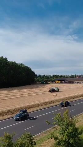 An aerial shot captures a rural scene. A vast field of golden hay stretches across the landscape, with rows of cut crops neatly arranged. A green tractor with a round bale attachment is actively working in the field, its presence suggesting agricultural activity. In the foreground, a paved road runs parallel to the field. A black car drives along the road from left to right. Further down the road, a white van travels in the same direction. To the left of the field, a dense line of dark green trees forms a natural border. In the distance, a cluster of buildings indicates a nearby town or settlement. The sky above is a vibrant blue, dotted with scattered white clouds.