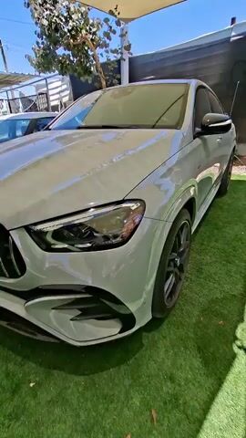 A side-view of a grey Mercedes-Benz GLE coupe is shown. The vehicle is parked on a patch of artificial grass. The camera pans from the front passenger side to the rear passenger side. The car has tinted windows and black rims. The sun is shining and casting shadows.
