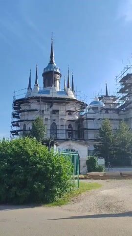 The video displays an exterior view of a large, multi-domed building undergoing extensive construction or renovation on a clear, sunny day. The camera slowly pans from left to right. The structure is predominantly white and features numerous turrets, spires, and domes, many of which are covered in extensive scaffolding. Some domes are visible and appear silver or light blue, while others are under construction. A dark-colored spire is prominent on the left side of the main building. In the foreground, a green metal gate is partially open, with green bushes and trees lining a dirt or gravel path. As the camera pans right, a separate, taller white structure, possibly a bell tower, with a light-colored dome is partially visible in the far right of the frame. No people are visible.