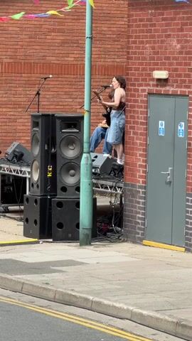 A woman stands on a platform, singing and playing a guitar. She is wearing a brown tank top and a denim skirt. Another person, wearing a blue shirt and jeans, is seated behind her. They are both performing on a makeshift stage constructed from speaker stacks and other equipment. The stage is set up against a brick building, with a green lamppost in the foreground. The area is outdoors, with a sidewalk and a street visible. Colorful banners are strung above the stage.