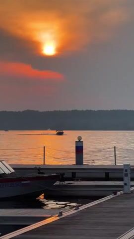 The video shows a sunset over a body of water. The sun is a bright orange ball low in the sky, surrounded by hazy clouds. A dark silhouette of a tree-covered shoreline is visible in the distance. In the foreground, a wooden dock extends into the water. A boat is docked on the left side of the frame, and another boat is moving across the water in the mid-ground, leaving a wake. A cylindrical marker with red, white, and blue bands stands on the dock. The water reflects the warm colors of the sunset. The overall mood is serene and tranquil.