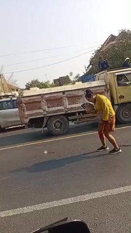 A woman wearing a yellow top and a patterned skirt walks on a paved road. She is near several large trucks, one of which is green and appears to be a dump truck. A silver car is also present. The woman makes a sudden movement downwards, appearing to stoop or crouch. She then stands up and continues walking. The environment appears to be outdoors, possibly during the day, with some vegetation and buildings in the background.