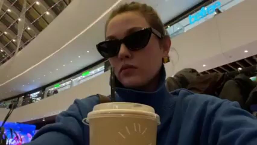 A young woman wearing sunglasses and a blue turtleneck sweater is sitting at a table in a shopping mall. She is drinking from a disposable cup with a straw. The cup has writing on it that includes the word "TIME." The background shows the mall's interior with shops and people. The woman appears to be looking directly at the camera.
