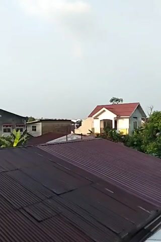 The video shows a panning view over a residential area with several houses and their roofs. The roofs are predominantly made of corrugated metal, with colors ranging from dark red to silver. In the background, a white house with a red roof is visible, surrounded by lush green trees. The sky is bright and overcast, suggesting it might be daytime. The overall scene depicts a typical urban or suburban neighborhood.