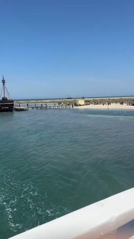 The video shows a wide shot of a calm, clear turquoise body of water in the foreground, with a shallow sandbar and beach visible in the background under a bright, cloudless blue sky. Several people are walking on a wooden pier extending into the water towards the sandbar. More people are scattered on the beach, some near small structures, possibly huts or umbrellas, suggesting a recreational area. A boat or ship is partially visible on the far left of the frame. The camera appears to be positioned on another boat, with a white railing in the lower right corner. The overall impression is a sunny day at a beach destination.