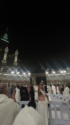 This video shows a nighttime scene at the Grand Mosque in Mecca. The Kaaba is visible in the background, illuminated by bright lights. A large crowd of people, many wearing ihram (white pilgrimage garments), are gathered around the Kaaba. In the foreground, a man in ihram is holding up his phone to take a selfie. He is smiling and appears to be capturing the moment. Other people are also visible in the crowd, some taking pictures and others engaged in prayer or conversation. The overall atmosphere is one of religious devotion and communal gathering. A clock tower is visible to the left of the frame.