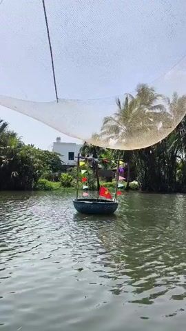 A small, round boat with a Vietnamese flag and colorful pennants is floating on a calm body of water. A large white net is suspended above the boat. The background includes lush green vegetation and palm trees. A white building is visible in the distance. The sky is bright.