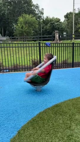 A young child is spinning around on a playground ride. The child is wearing a red and green shirt and green shorts. The ride is a large, dark gray and blue pod that is attached to a central pole. The pod is spinning rapidly, creating a blur of motion. The playground surface is blue and green, with a black metal fence in the background. Trees and a cloudy sky are visible beyond the fence.