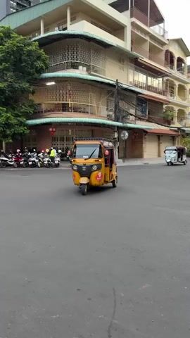 A yellow tuk-tuk is driving down a street in an urban environment. The tuk-tuk is the primary focus of the video. In the background, there are several buildings, other vehicles including motorcycles and another tuk-tuk, and some trees. The street appears to be paved and is wet, suggesting it might have recently rained. The time of day appears to be either early morning or late afternoon, as the lighting is somewhat dim. The video is shot from a slightly low angle, looking up at the tuk-tuk. The tuk-tuk has a license plate that reads "P2636". There are also some logos and stickers on the exterior of the tuk-tuk. The interior of the tuk-tuk is visible, showing seats and some equipment. There are no people clearly visible inside or around the tuk-tuk, though a person is partially visible driving it. The video is 3 seconds long.