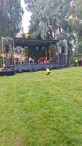 The video opens with a shot of an outdoor stage set up on a grassy area, with trees in the background. There are several people visible on the stage, appearing to be setting up equipment or preparing for a performance. In the foreground, a person wearing a yellow vest and dark pants is seen jogging across the grass. As the video progresses, more people come into view, some of whom appear to be dancing or moving energetically on the grass. There is a group of individuals on the stage, and at least two people in the foreground are engaged in some form of physical activity. The lighting suggests it is either early morning or late afternoon.